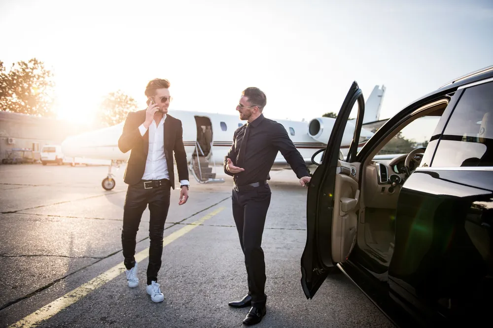 Man on phone exiting car at airfield with private jet in background.