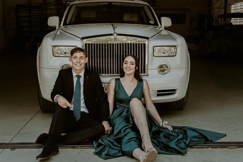 Couple sitting in front of white Rolls-Royce. Man in suit, woman in emerald gown, smiling. In a garage.