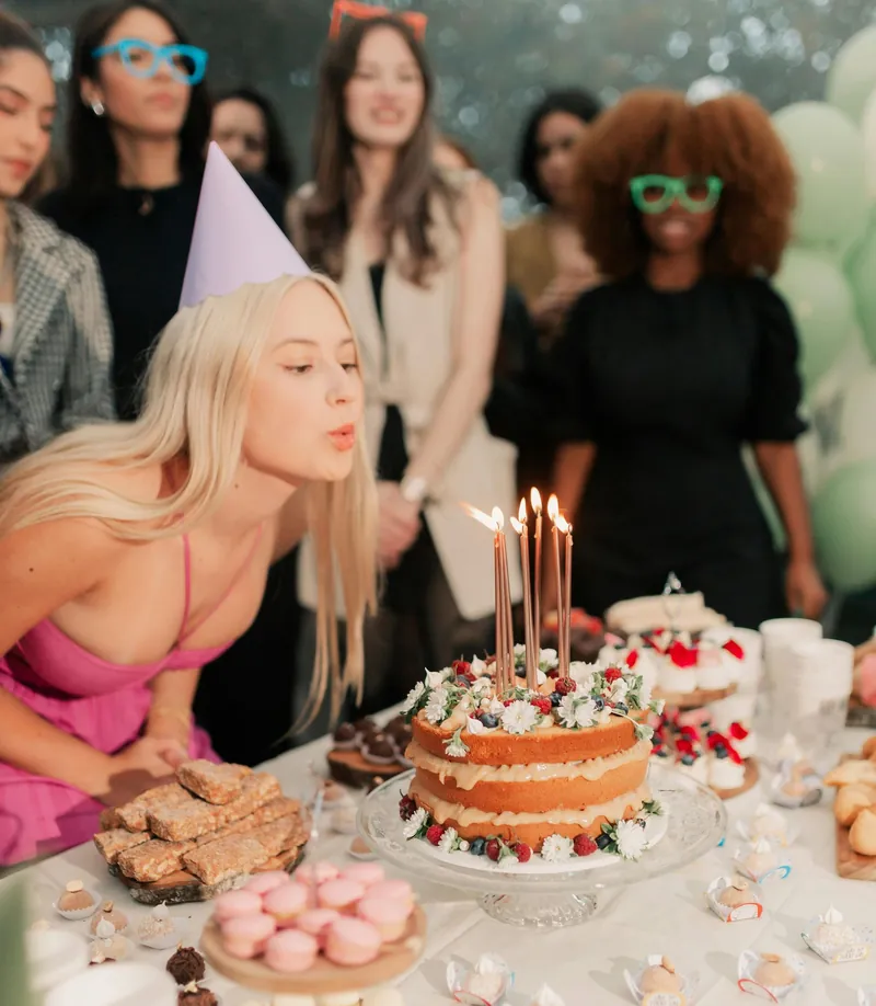 Blonde woman in party hat blows out candles on cake at birthday party with friends.
