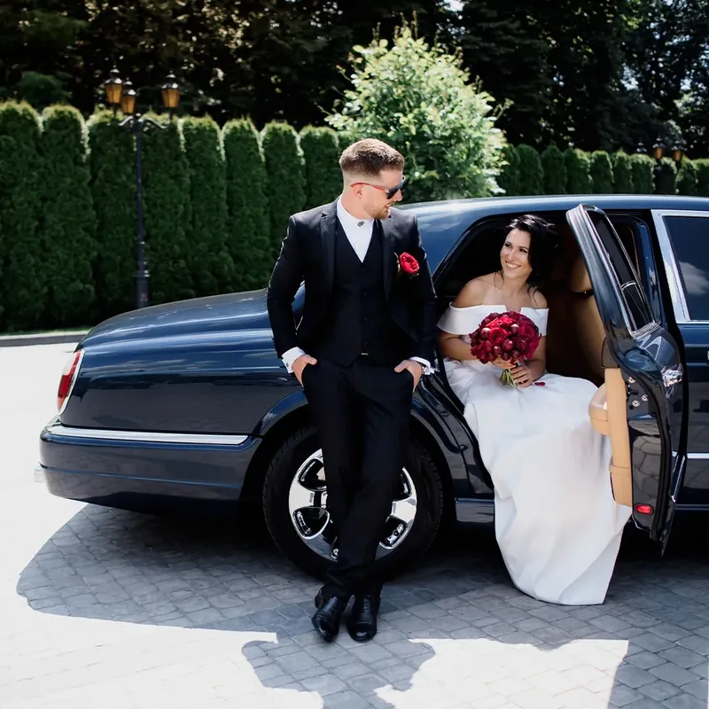 A newlywed couple by a black limousine. The bride in a white gown holds a bouquet. 